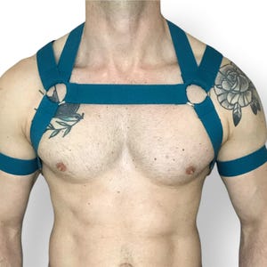 Puede incluir: Arnés corporal de color turquesa con anillos metálicos. El arnés presenta correas en el pecho, los hombros y los brazos. El diseño es sencillo y moderno, adecuado para moda o fetiche.