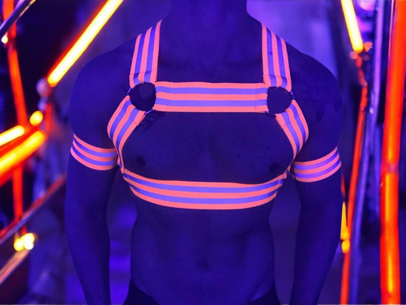 Made-to-measure men’s elastic chest harness in high-visibility neon orange with reflective detailing, glowing under blacklight. The design features double shoulder and chest straps joined by circular O-rings, with optional matching jockstrap and thigh bands—ideal for clubwear, festivals, or fetish fashion.