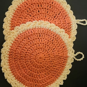 May include: Two crocheted pot holders with a peach colored center and a cream colored border. The pot holders have a loop handle.