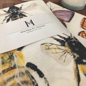 Puede incluir: Dos paños de cocina blancos con ilustraciones de acuarela de abejas y mariposas. Los paños están envueltos con una banda blanca que dice "Meg Hayes Art - Tea Towel" y "meghayesart.co.uk". Los paños son de tela clara.