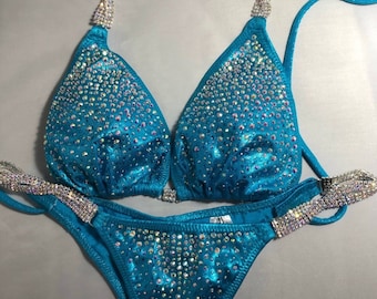 May include: A turquoise blue bikini with crystal embellishments. The bikini top has a halter style with a strap that wraps around the neck. The bikini bottom has a high-cut style.