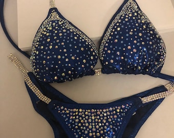 May include: A navy blue bikini with crystal embellishments. The bikini top has a halter style with a strap that goes around the neck and ties at the back. The bikini bottom has a high-cut style.