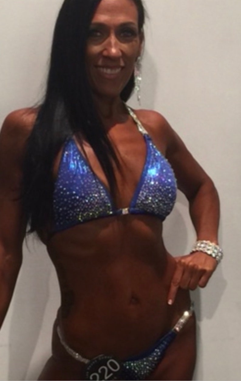May include: A woman wearing a blue and silver sequined bikini with rhinestones. She is posing for a competition and has a number "220" on her bikini bottom.