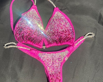 Può includere: Un bikini rosa con decorazioni di cristalli. La parte superiore ha uno stile halter e la parte inferiore è un taglio audace.