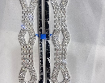 May include: Two silver-toned rhinestone shoe clips, each measuring approximately 5.5 inches in length. The clips feature a repeating pattern of ovals and squares, with a loop at the top and bottom.