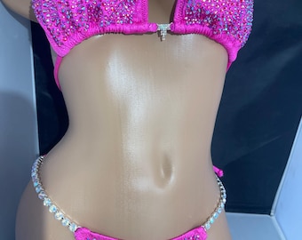 Può includere: Un bikini rosa con decorazioni di cristalli. La parte superiore ha una scollatura profonda e la parte inferiore è un taglio audace.