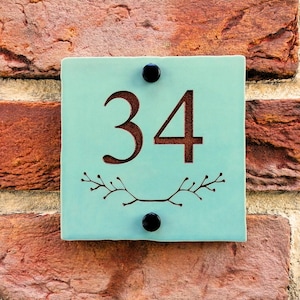 May include: A turquoise blue square house number sign with black screws and a brown number "34" on a brick wall.