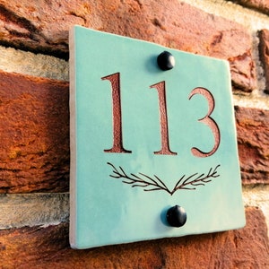 Könnte beinhalten: Ein türkisblaues Keramik-Hausnummernschild mit der Nummer 113 in brauner Schrift. Das Schild ist mit zwei schwarzen Schrauben an einer Backsteinwand befestigt.