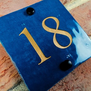 May include: A blue square house number sign with gold numbers "18" on it. The sign is attached to a brick wall with black screws.