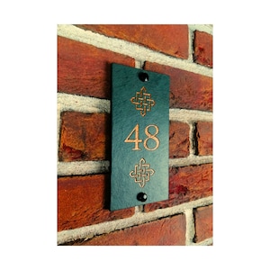 Puede incluir: Un letrero de número de casa de pizarra verde oscuro con un número 48 de color cobre y dos diseños de nudos celtas. El letrero está montado en una pared de ladrillo.