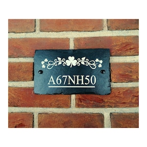 Puede incluir: Un letrero rectangular de pizarra negra para casa con texto blanco y elementos florales decorativos. El letrero muestra la dirección "A67NH50" y está montado en una pared de ladrillo rojo. Dos tornillos negros aseguran el letrero.