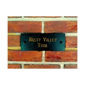 Puede incluir: Un letrero de pizarra negro con letras doradas que dice "Misty Valley Trail" montado en una pared de ladrillo.