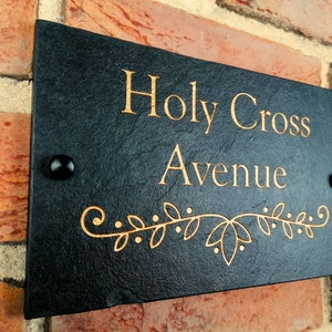 Könnte beinhalten: Schwarzes Schiefer-Hausnummernschild mit goldener Beschriftung "Holy Cross Avenue" und einem dekorativen goldenen Blumenmuster.