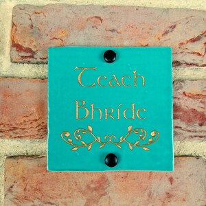 May include: Teal blue square sign with gold lettering that reads "Teach Bhride" with a gold decorative vine design at the bottom. The sign is attached to a brick wall with two black tacks.