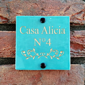 以下が含まれることがあります： 「Casa Alicia N°4」と金色で書かれたターコイズブルーのセラミック製の家の番号札。札には装飾的な花の模様と黒いネジが2つ付いている。
