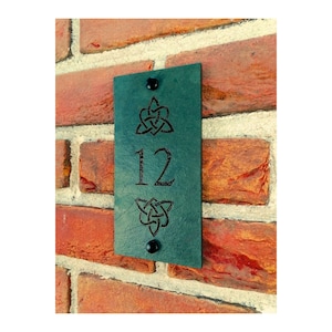 May include: A teal rectangular house number sign with the number 12 in the center. The sign has two black fasteners and Celtic knot designs above and below the number. The sign is mounted on a red brick wall.