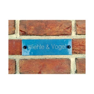 May include: A blue glass sign with the text "Wiehle & Vogel" engraved in white on a brick wall.