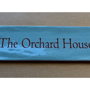 May include: A light blue ceramic house sign with the text "The Orchard House" in red lettering.