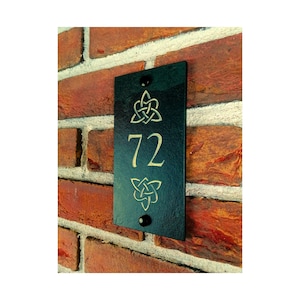 May include: Black rectangular address plaque with gold-colored Celtic knot designs and the number 72. The plaque is mounted on a red brick wall with two black screws.