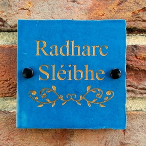 May include: A blue slate sign with gold lettering that reads "Radharc Sléibhe". The sign has two black screws on either side and a decorative gold vine design at the bottom.