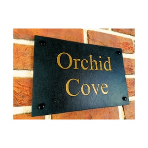 Puede incluir: Un letrero de casa de pizarra negra con letras doradas que dice "Orchid Cove".