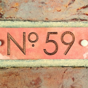 May include: A pink ceramic house number plaque with the number "No. 59" in brown lettering. The plaque is attached to a brick wall with two white round fasteners.