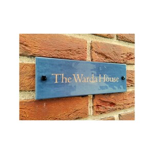 以下が含まれることがあります： 金色の文字で「The Warda House」と書かれたスレート製の家の看板。看板は2本の黒いネジでレンガの壁に取り付けられています。