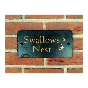 Peut inclure: Une plaque de maison en ardoise noire avec une inscription dorée "Swallows Nest" et une silhouette d'hirondelle dorée à droite. La plaque est fixée à un mur de briques.