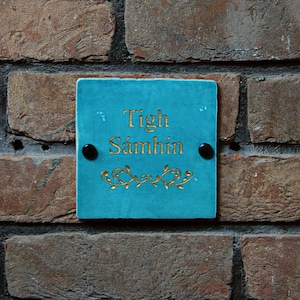 Op de afbeelding: Een tealblauw bord met gouden letters met de tekst "Tigh Samhain" op een bakstenen muur.