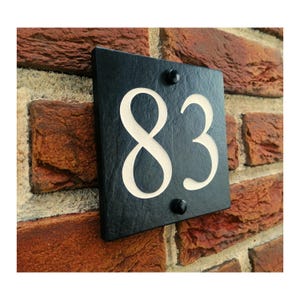 May include: A black slate house number plaque displaying the number 83 in a cream-coloured, stylised font. The square plaque is mounted on a red brick wall with two black screws. The numbers are carved out, revealing the cream-coloured backing.