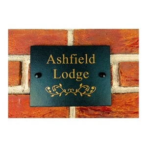 Könnte beinhalten: Schwarzes Schiefer-Hausschild mit goldener Beschriftung "Ashfield Lodge" und einem goldenen dekorativen Rankenmuster.