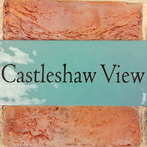 以下が含まれることがあります： 「Castleshaw View」と黒文字で書かれた、ターコイズブルーの長方形の家の番号札。レンガの壁に取り付けられています。