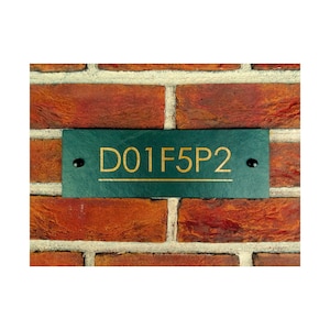 Puede incluir: Un letrero de número de casa de pizarra verde con letras doradas que dice "D01F5P2" montado en una pared de ladrillo.