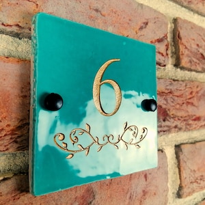 Puede incluir: Placa de número de casa cuadrada de color azul turquesa con el número 6 en dorado y un diseño floral dorado. Se ven dos tornillos negros en la placa.