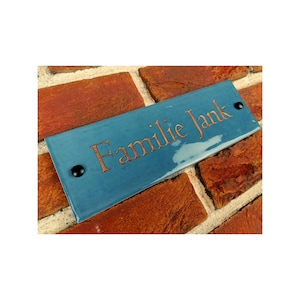 May include: A blue ceramic house number sign with the text "Familie Jank" in copper lettering. The sign is mounted on a brick wall.