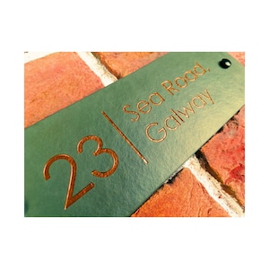 Puede incluir: Un letrero rectangular de color verde azulado con el número "23" y el texto "Sea Road, Galway" en letras cobrizas. El letrero tiene un pequeño agujero en la parte superior y está sobre un fondo de ladrillos.