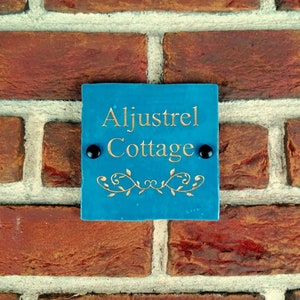 May include: A blue square sign with gold lettering that reads "Aljustrel Cottage". The sign has a decorative gold vine design and is attached to a brick wall with two black screws.