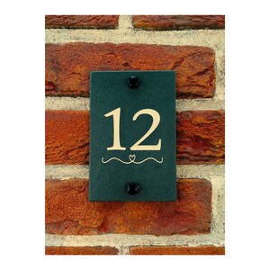 Puede incluir: Un letrero de número de casa de pizarra verde oscuro con el número 12 en color crema. El letrero está fijado a una pared de ladrillo.