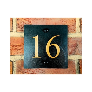 Puede incluir: Placa de número de casa de pizarra negra con el número 16 pintado en oro. La placa está montada en una pared de ladrillo.