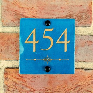 May include: A blue square tile with gold numbers 454 and a decorative design. The tile is mounted on a brick wall.