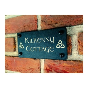Op de afbeelding: Een zwart leisteen huisbord met gouden letters met de tekst "Kilkenny Cottage" en een Keltisch knoopontwerp aan beide zijden.