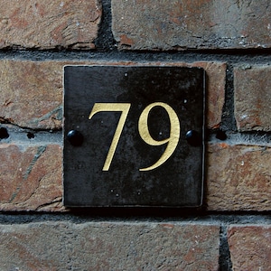 Könnte beinhalten: Ein schwarzes quadratisches Schild mit goldenen Zahlen "79" an einer Backsteinwand.
