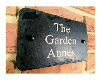 May include: A rectangular black slate sign with the words "The Garden Annex" in a cream-colored font. The sign is mounted on a brick wall and has two black mounting screws.