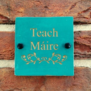 May include: A square, teal-colored sign with the words "Teach Máire" in gold lettering. The sign is adorned with a gold floral design and is mounted on a brick wall with two black screws.