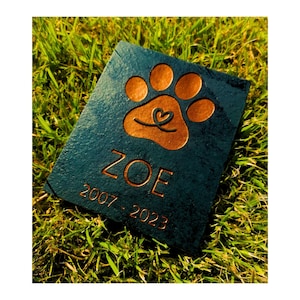 May include: A dark blue slate pet memorial with a copper-coloured paw print and heart design. The name "ZOE" and the dates "2007-2023" are engraved on the stone.
