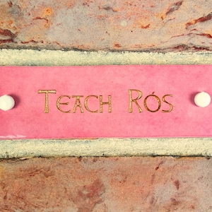 May include: A pink ceramic sign with white dots on either side and the text "Teach Rós" in gold lettering.