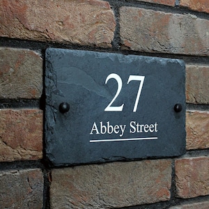 May include: Black slate house number sign with white lettering. The number 27 and the street name Abbey Street are displayed on the sign.