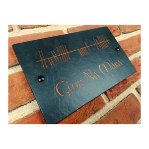 Puede incluir: Un letrero rectangular de pizarra negra con texto y símbolos grabados en color cobre. El texto dice "Glór Na Mara" en una fuente estilizada. Dos remaches negros aseguran el letrero. El letrero está montado en una pared de ladrillo rojo.