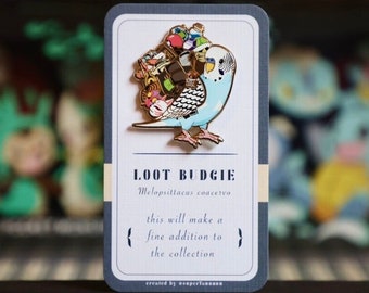May include: A blue and white Loot Budgie enamel pin with a gold outline, carrying a pile of colorful items. The pin is on a card with the text "LOOT BUDGIE" and "this will make a fine addition to the collection."