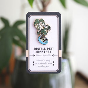 May include: A gold-tone enamel pin featuring a Monstera deliciosa plant growing out of a blue and green digital pet toy. The pin has the text "Digital Pet Monstera" and "Monstera digitusdelicia" on it. The text "where we're going, we won't need a green thumb to grow" is also on the pin.
